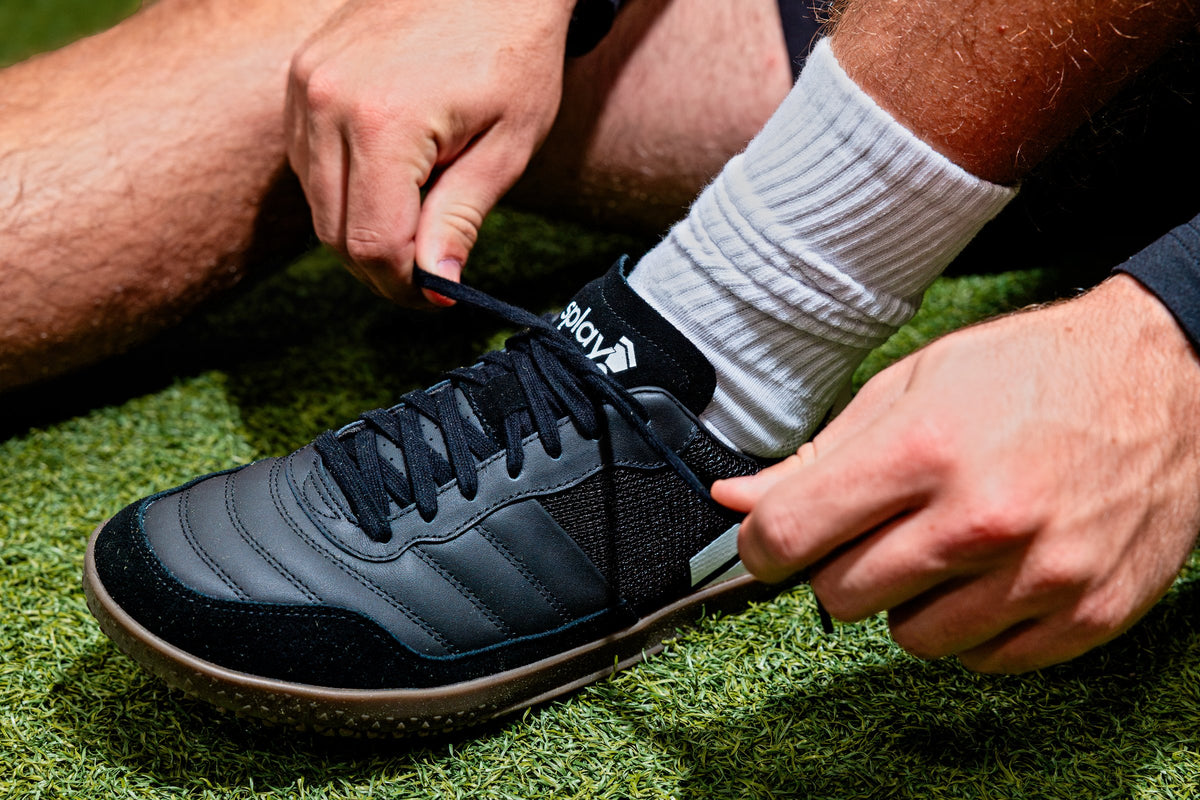 Close-up of hands lacing a black Dominari Indoor shoe on turf