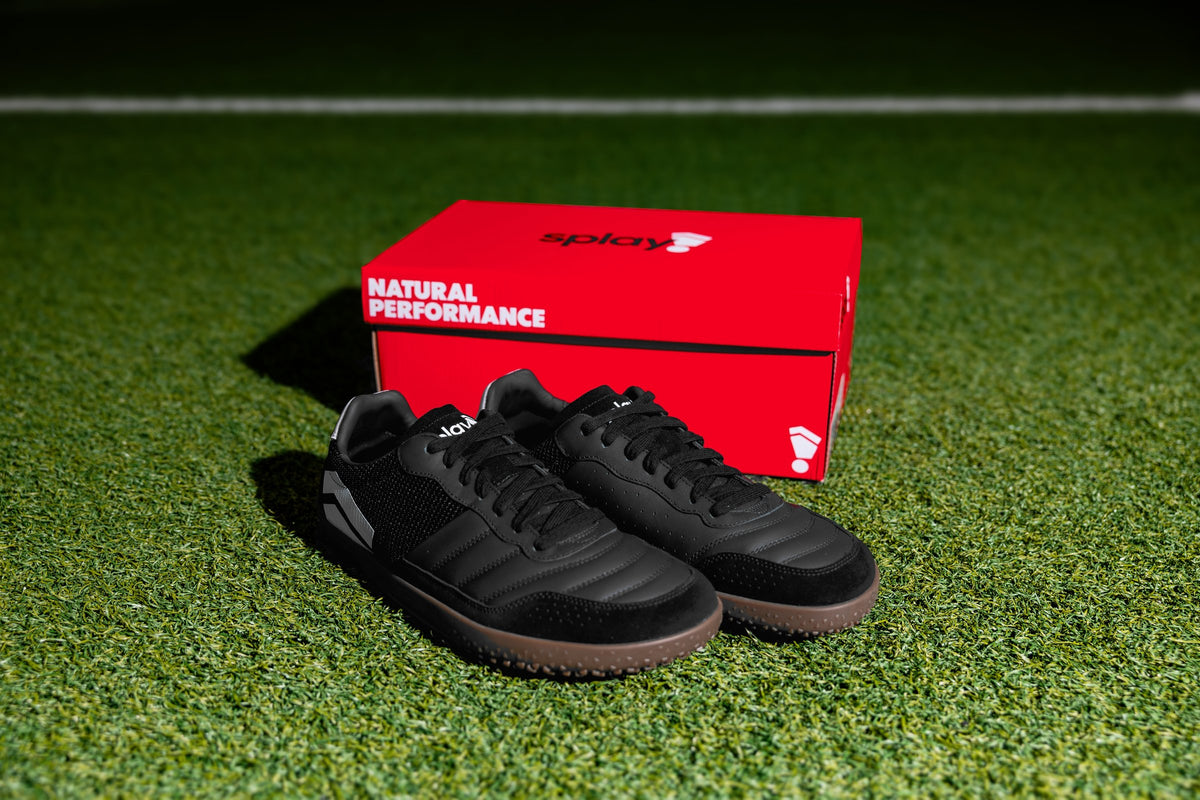 Splay Dominari Indoor shoes in black with gum sole beside a red Splay box on turf
