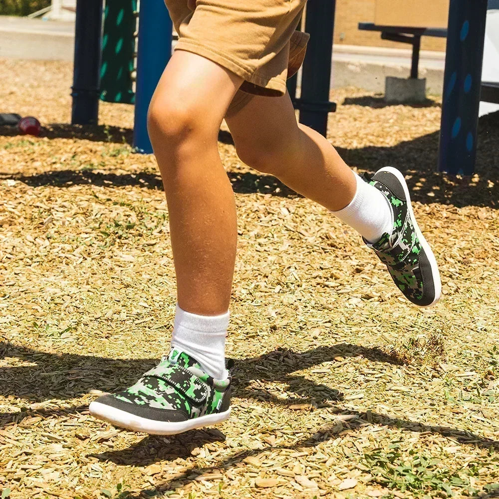 Child running across a playground wearing green camouflage Splay barefoot shoes with hook-and-loop straps, white socks, and gray shorts.