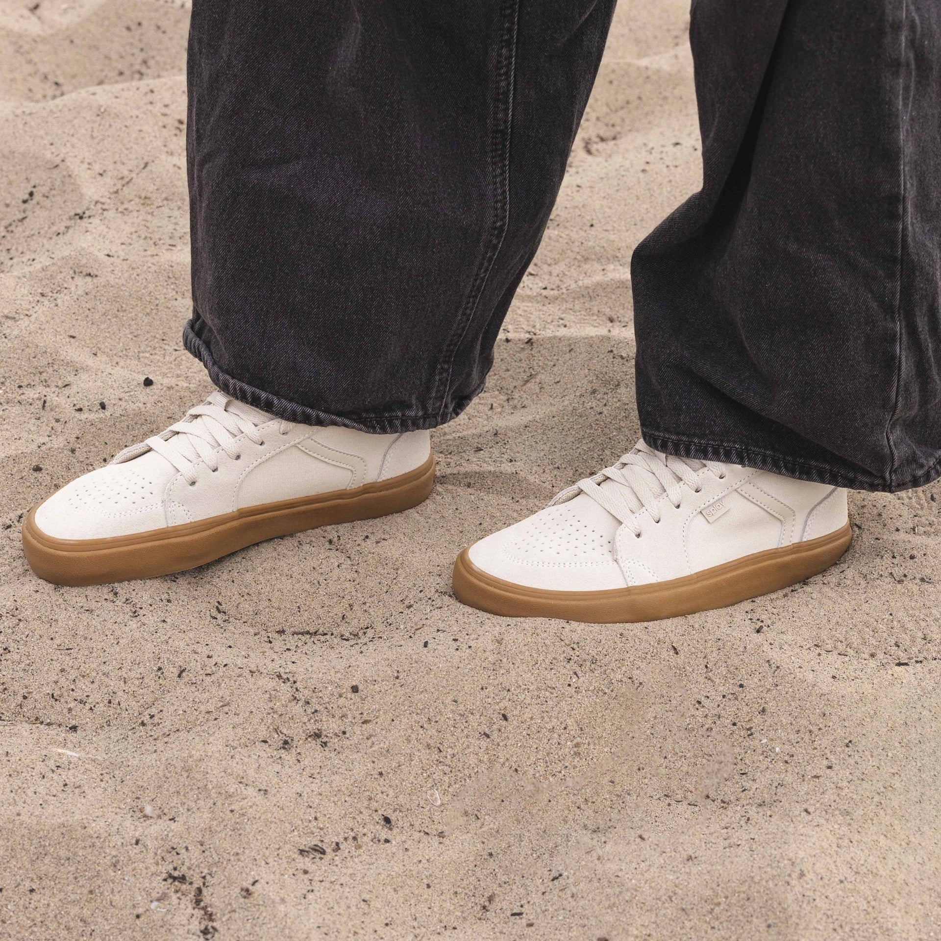 Person standing on sand wearing white Splay barefoot sneakers with gum soles and black jeans.