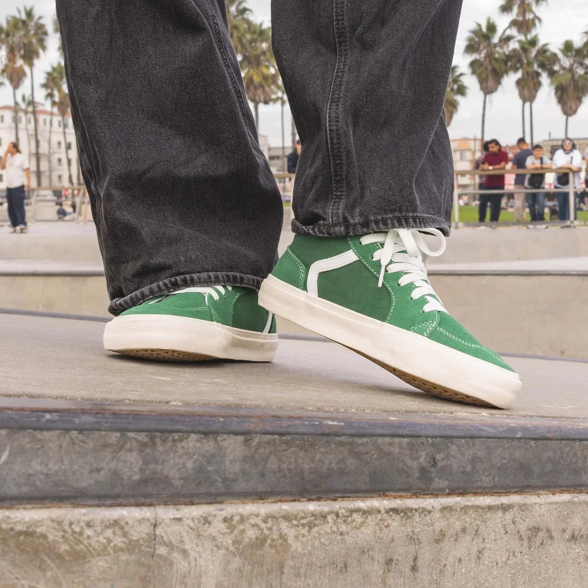Person wearing green Splay barefoot sneakers with white laces and side stripes, standing on a concrete ledge at a skatepark with palm trees in the background.