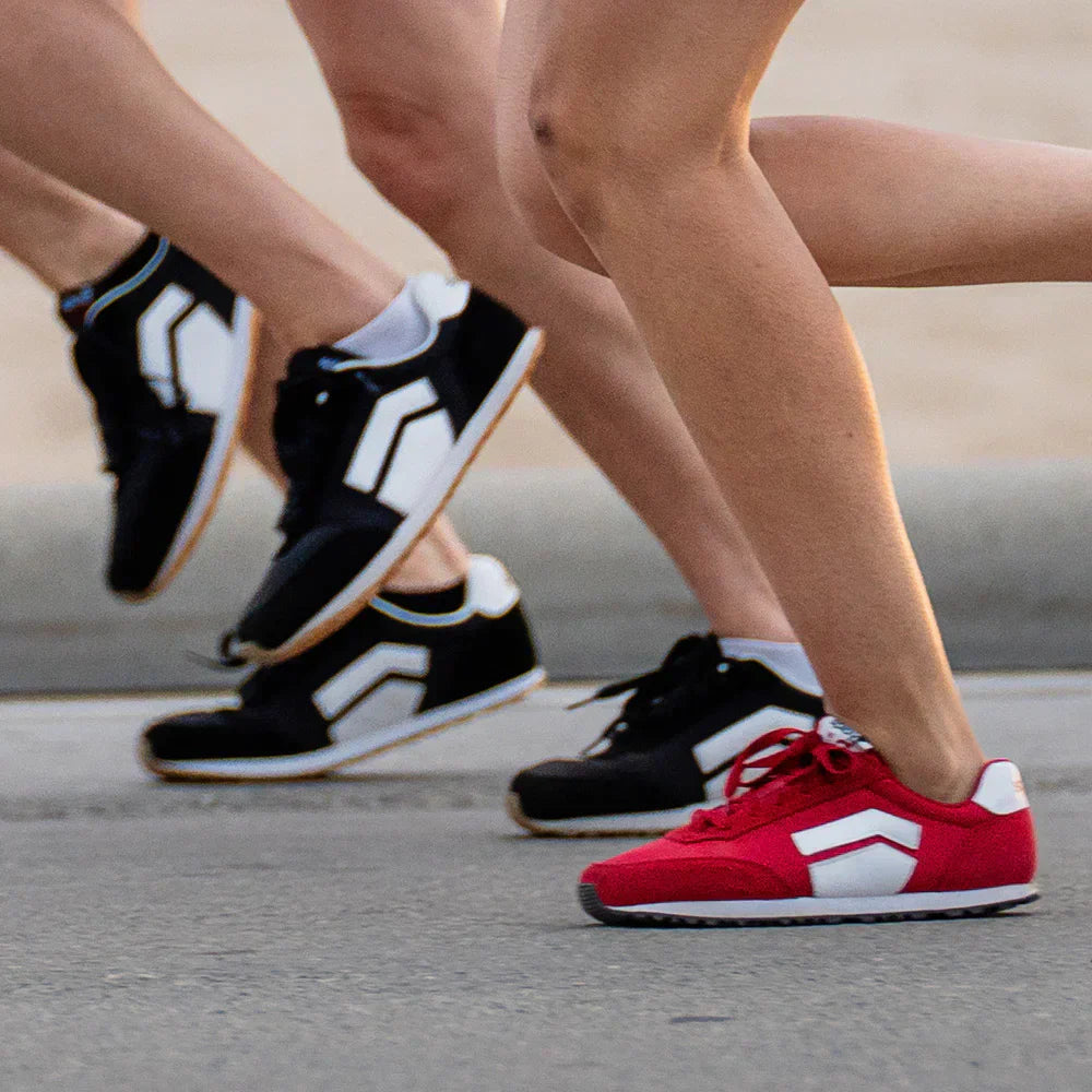 Two runners mid-stride on pavement wearing Splay RUNNER V1 zero-drop shoes in black and red colorways.