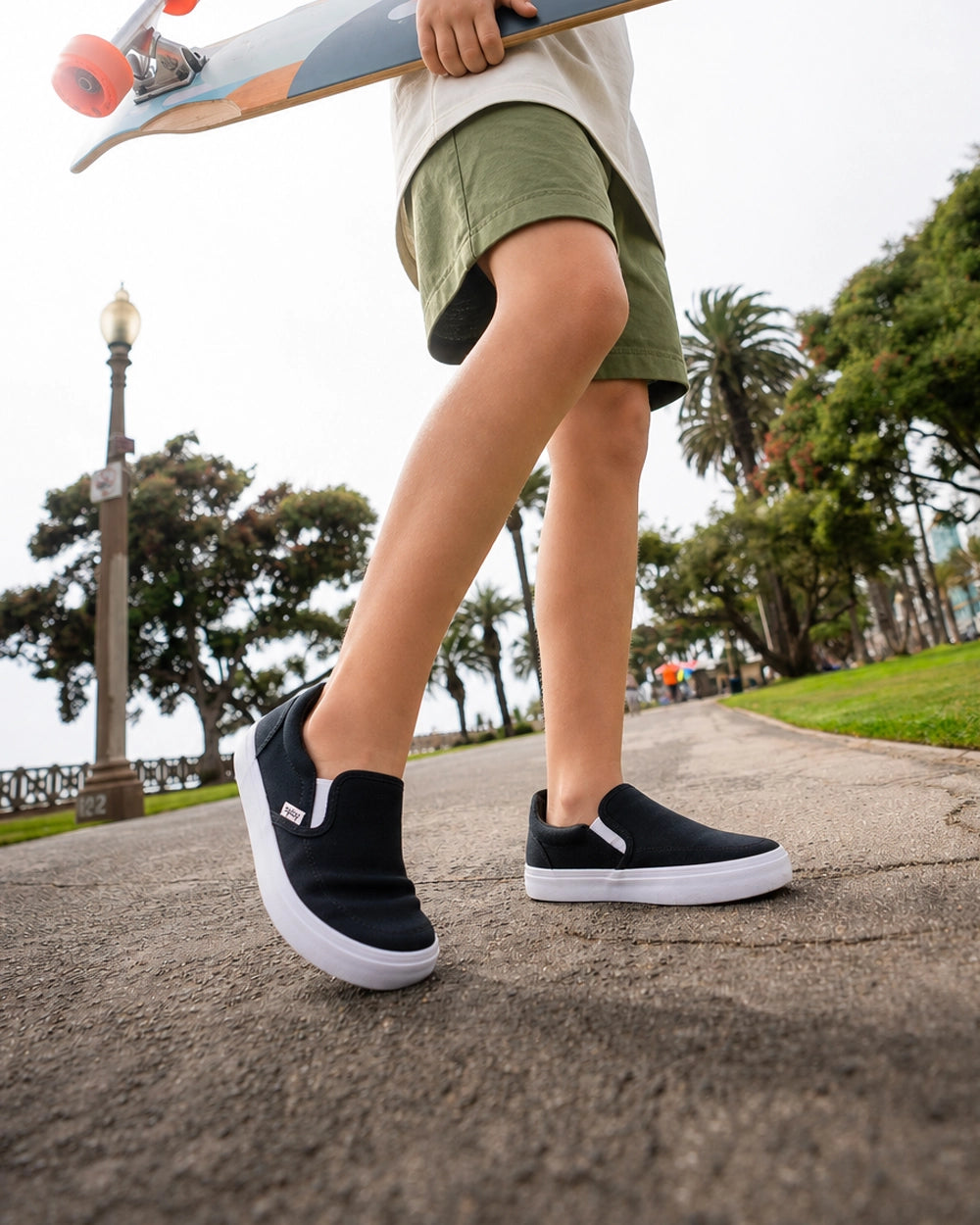 Child wearing black Splay Kids REV SO Berlin barefoot slip-on shoes with white soles while carrying a skateboard outdoors.