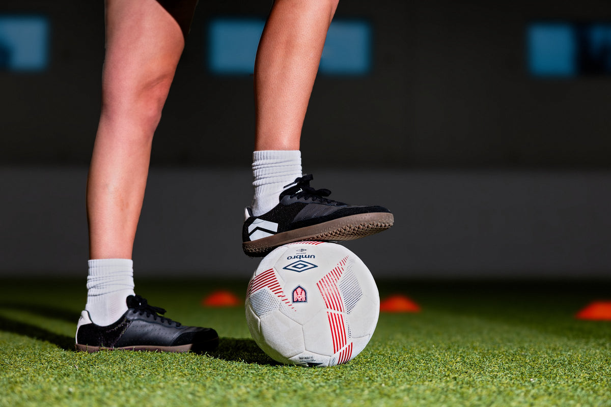 Player resting a Dominari Indoor shoe on a soccer ball on indoor turf