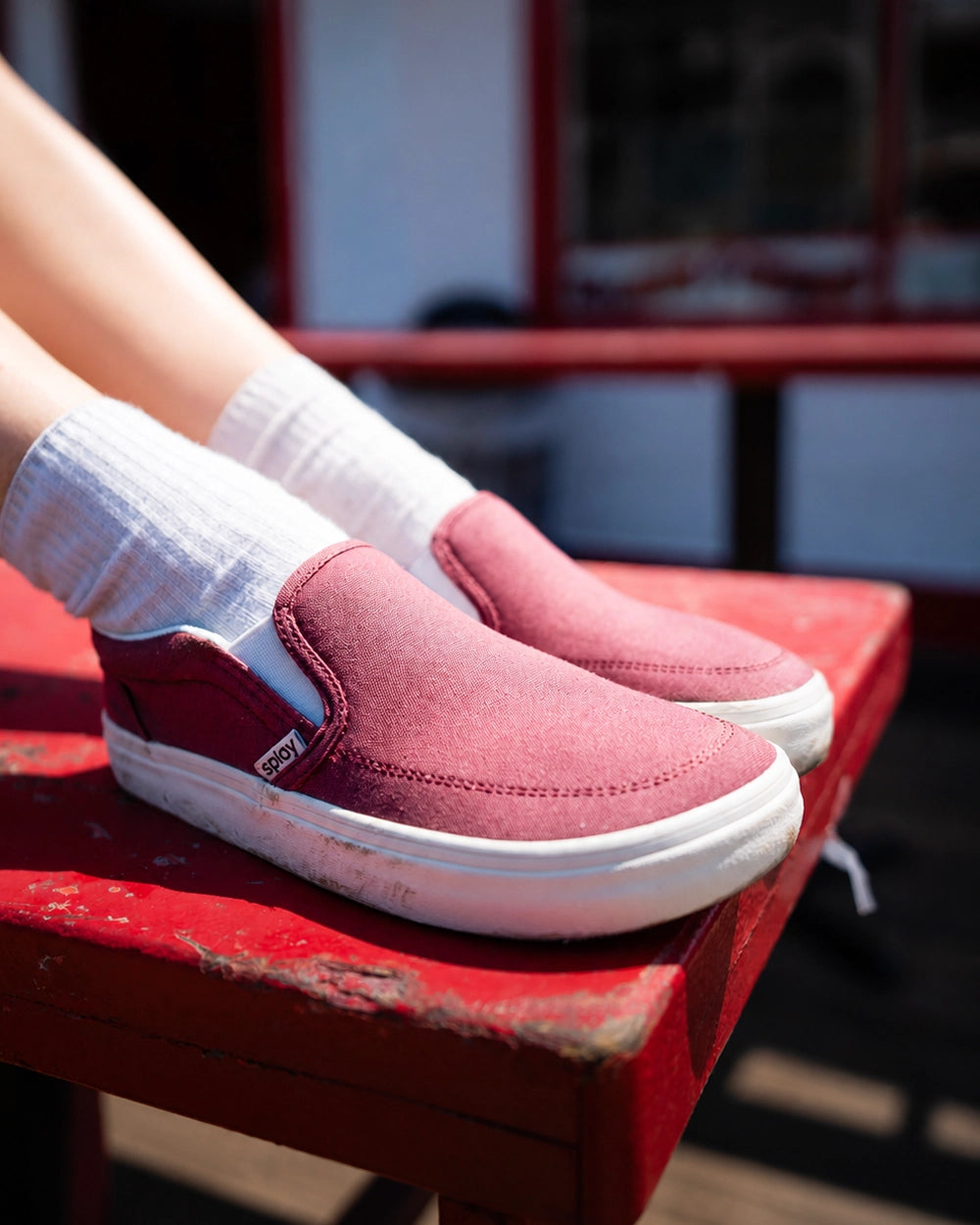 Child wearing burgundy Splay Kids REV SO Madrid barefoot slip-on shoes with white socks, resting their feet on a red bench outdoors.