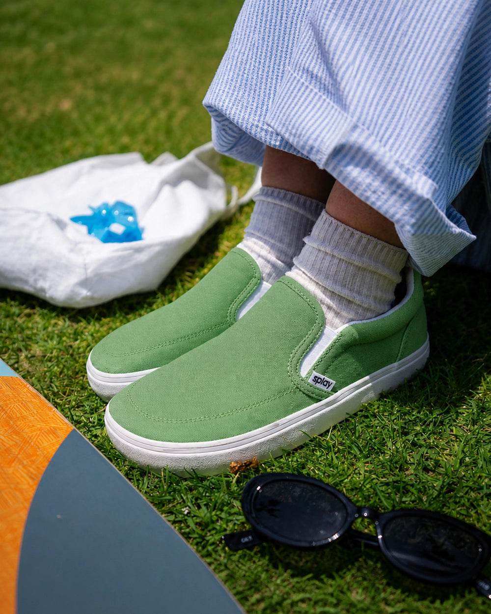 Child wearing green Splay Kids REV SO Rio barefoot slip-on shoes while seated on grass beside a skateboard and sunglasses.